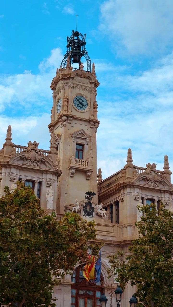 Torre dell'orologio del Municipio di Valencia sotto un cielo blu nuvoloso. Vista dettagliata della torre dell'orologio ornata e della facciata dell'edificio dell'Ayuntamiento de Valencia con bandiere spagnole.