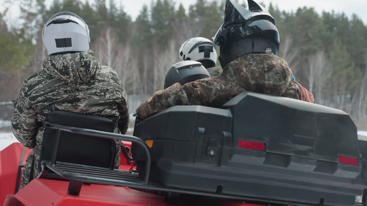 Escursione invernale in quad attraverso una foresta innevata, un quad rosso trasporta piloti ben coperti con giacche mimetiche e caschi; la guida e il conducente si preparano al punto di partenza, il motore romba, i pneumatici sollevano la neve lungo il sentiero fiancheggiato da pini.