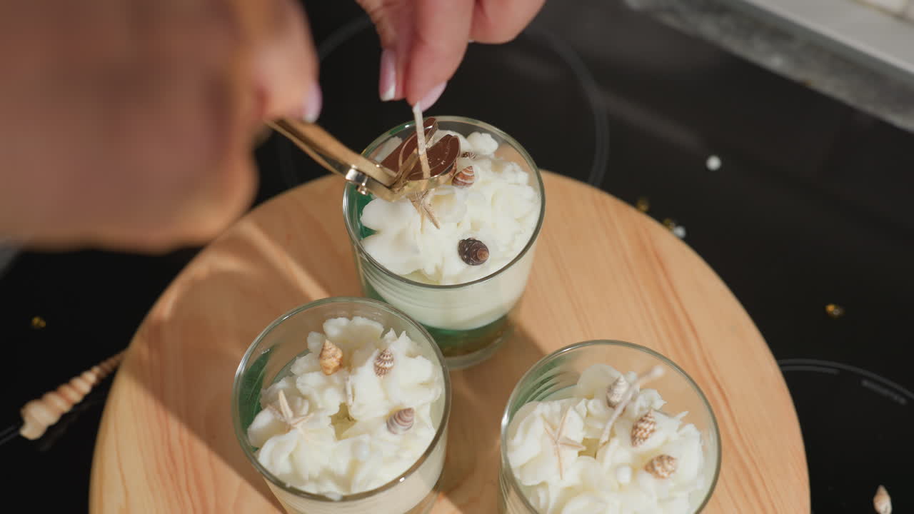 House wife trims long wick in center candle jar using golden wick trimmer while holding it steadily, surrounded by two more decorative wax jars on wooden tray adorned with miniature shells