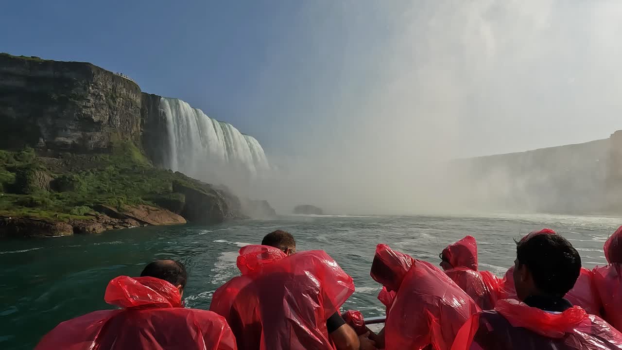 Tourists in red ponchos onboard a boat approaching Niagara Falls, with mist rising and lush cliffs under a sunny sky, Canada