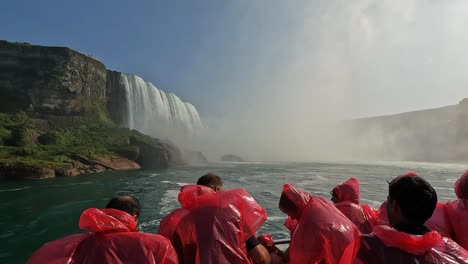 tourists in red ponchos onboard a boat approaching niagara falls, with mist rising and lush cliffs under a sunny sky, canada
