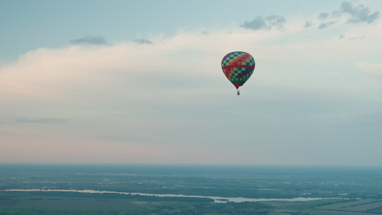 close up of videographer capturing sprawling green farmland from aerial vantage as distant multicolored hot air balloon drifts gracefully over patchwork fields under clear blue sky at golden hour
