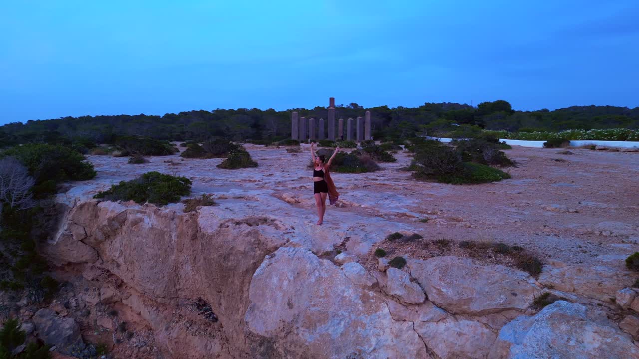 Woman performing a yoga tree pose on a rocky cliff during a serene sunset in Cala Llentia Ibiza. Amazing aerial view flight panorama orbit drone
