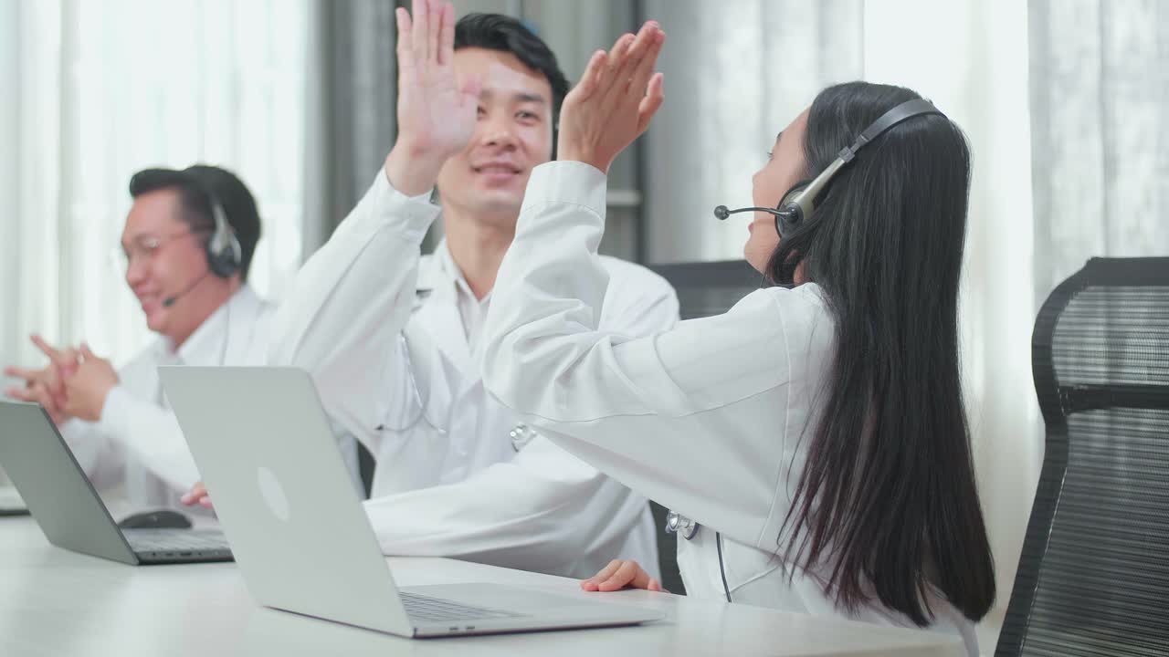 Three Asian Doctors With Stethoscopes In Headsets Working As Call Center Agents Sit On The Chairs And And Being Happy Due To Success Working With Computers At The Office