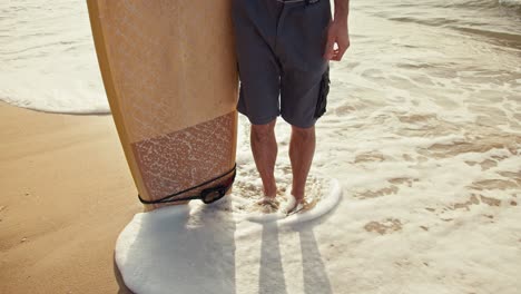 Close-up-shot-from-above,-a-guy-in-blue-shorts-holds-a-yellow-wooden-textured-surfboard-and-stands-on-orange-sand-on-the-seashore-whose-waves-come-to-the-guy’s-feet-and-wrap-around-them-in-the-morning