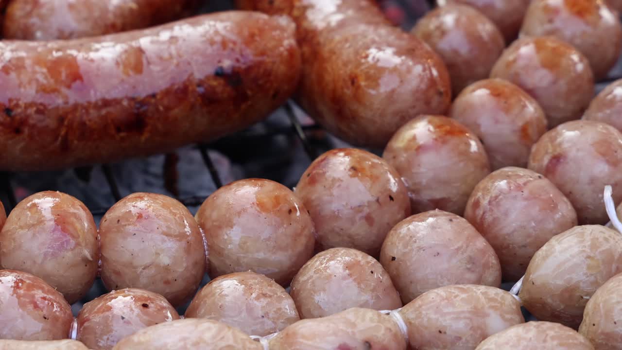 Close-up of sausages and meatballs grilling over open flame, glistening with oil and heat