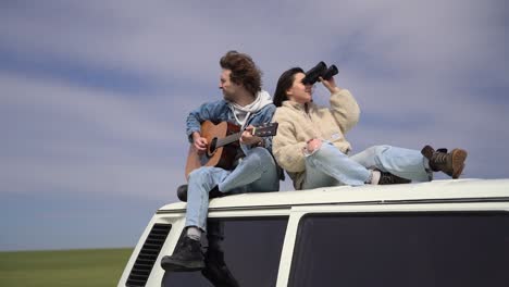 a young boy plays the guitar and a young girl looks around with a pair of binoculars on the roof of a caravan.