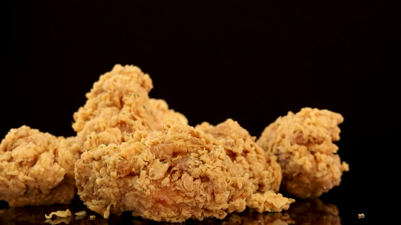 Golden, crumbed chicken breasts and thighs rotate slowly on a reflective surface with dramatic studio lighting and a dark background, highlighting texture and seasoning