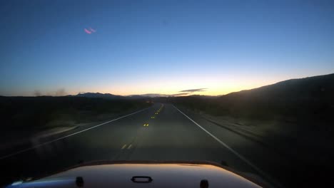 driving toward the sunrise in a red vehicle in the desert with light from headlights and mountains visible in silhouette