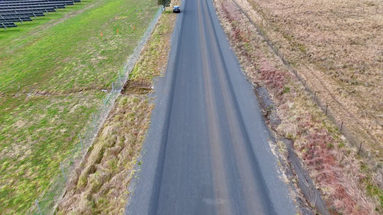 A single vehicle travels along an empty country road bordered by grass and fields, captured in daylight from a high, overhead drone perspective