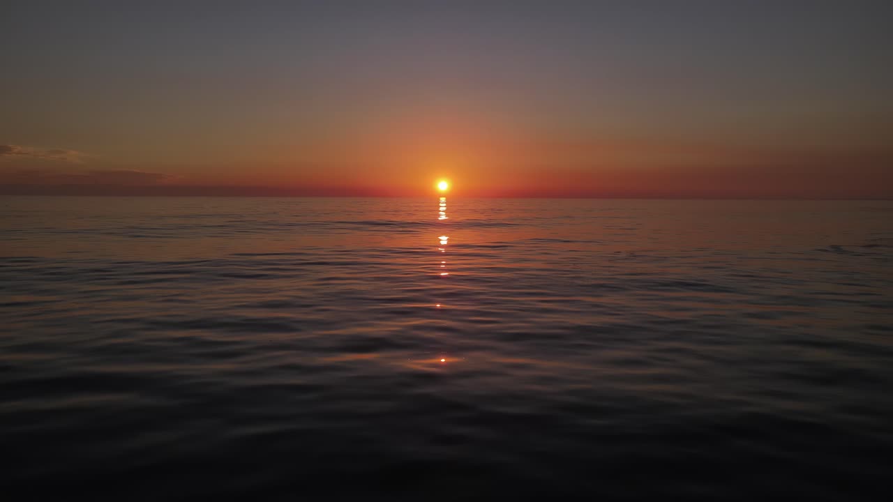 중앙에 황금빛 태양이 지는 잔잔한 바다 위를 날아가는 드론 샷