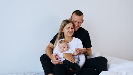 young caucasian couple sit with their baby in hands. dad holds the tiny kid's hand. white backdrop.
