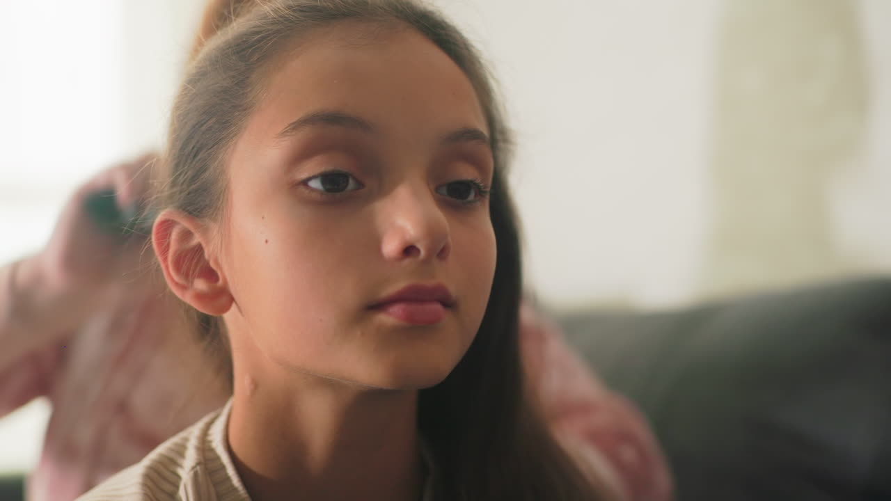 Mother brushing daughter’s hair in cozy living room. Close-up of daughter’s face as she relaxes, showing calm and peaceful moment. Soft natural light filtering through window, creating warm atmosphere