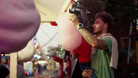 a little blond boy in a red t-shirt takes pink cotton candy from a sweet shop in an amusement park with his dad. a little boy in a red t-shirt sits in the arms of his dad in a green t-shirt and eats cotton candy