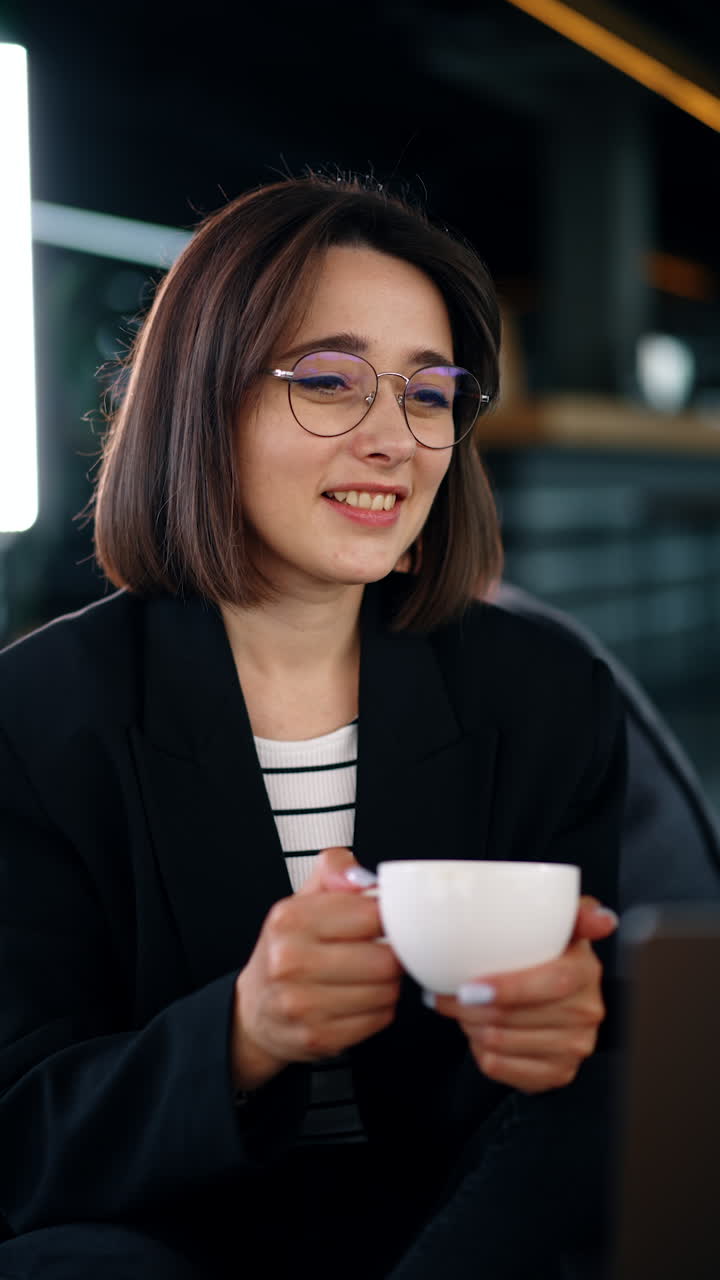 Beautiful Caucasian brunette woman wearing glasses holding a cup. Smiling lady looks at her laptop. Vertical video.