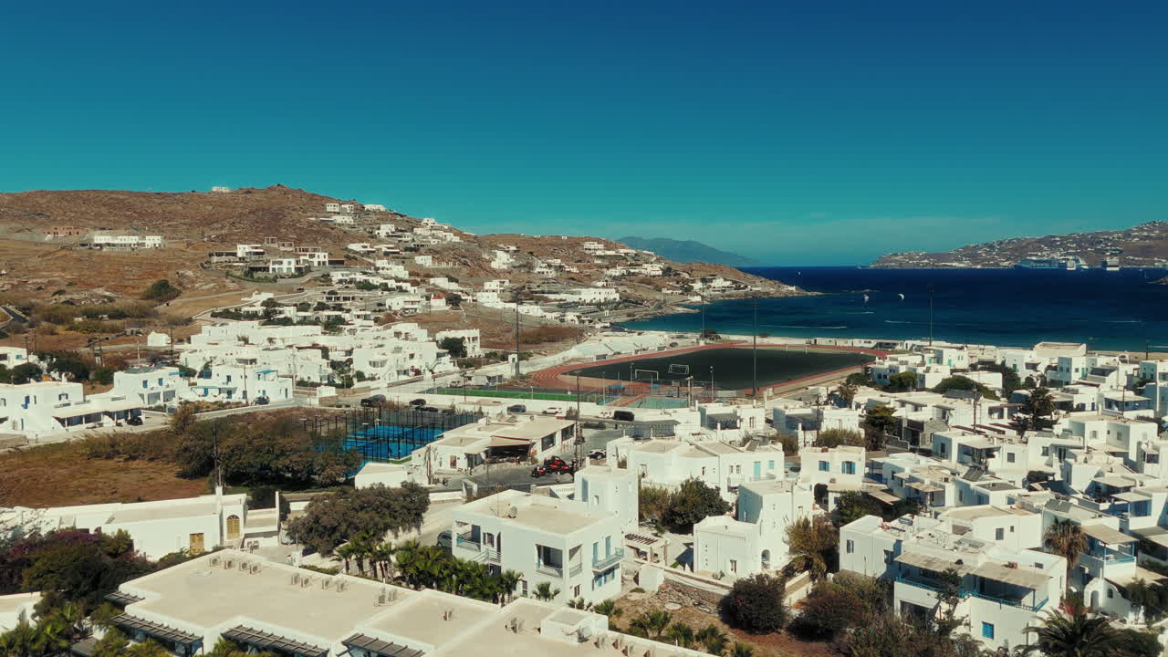 Drone shot moving over Mykonos town showing white houses, sports field, and the blue Aegean Sea