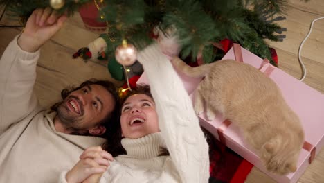 Close-up-shot,-a-guy-and-a-brunette-girl-in-a-white-sweater-are-lying-on-the-floor-on-a-red-blanket-and-looking-at-a-green-decorated-Christmas-tree-near-their-cream-colored-cat-in-a-cozy-room