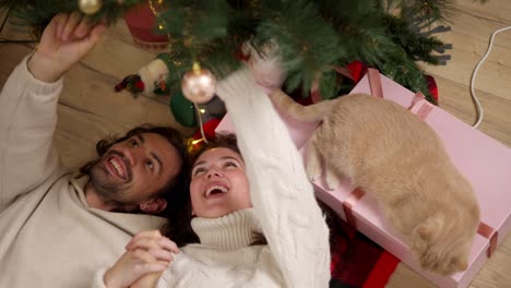 en primer plano, un chico y una chica morena con un suéter blanco están tendidos en el suelo sobre una manta roja y mirando un árbol de navidad decorado de verde cerca de su gato de color crema en una habitación acogedora