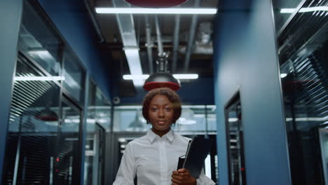 Cheerful-afro-woman-looking-at-camera-in-corridor.-Woman-smiling-in-office