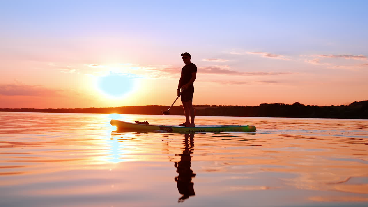 Sportsman slowly rows with an oar floating by the river. Man goes sup boarding at sunset. Side low angle view.