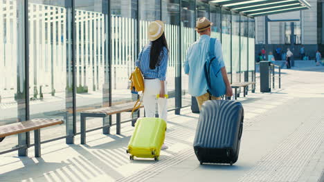 Rear-view-of-caucasian-young-couple-of-travellers-wearing-hat-with-backpack-and-carrying-suitcases-in-the-bus-stop