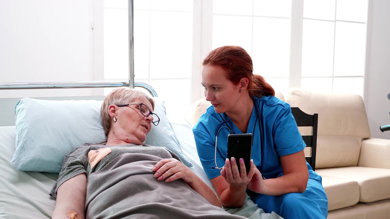 Nurse Showing Smartphone to Elderly Patient in Hospital Bed