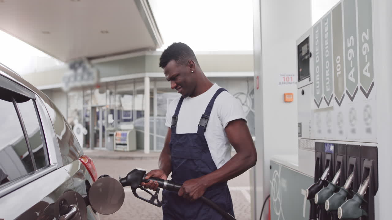 Pæn ung afrikansk mandlig tankstationsmedarbejder iført hvid t-shirt og blå overalls og holder