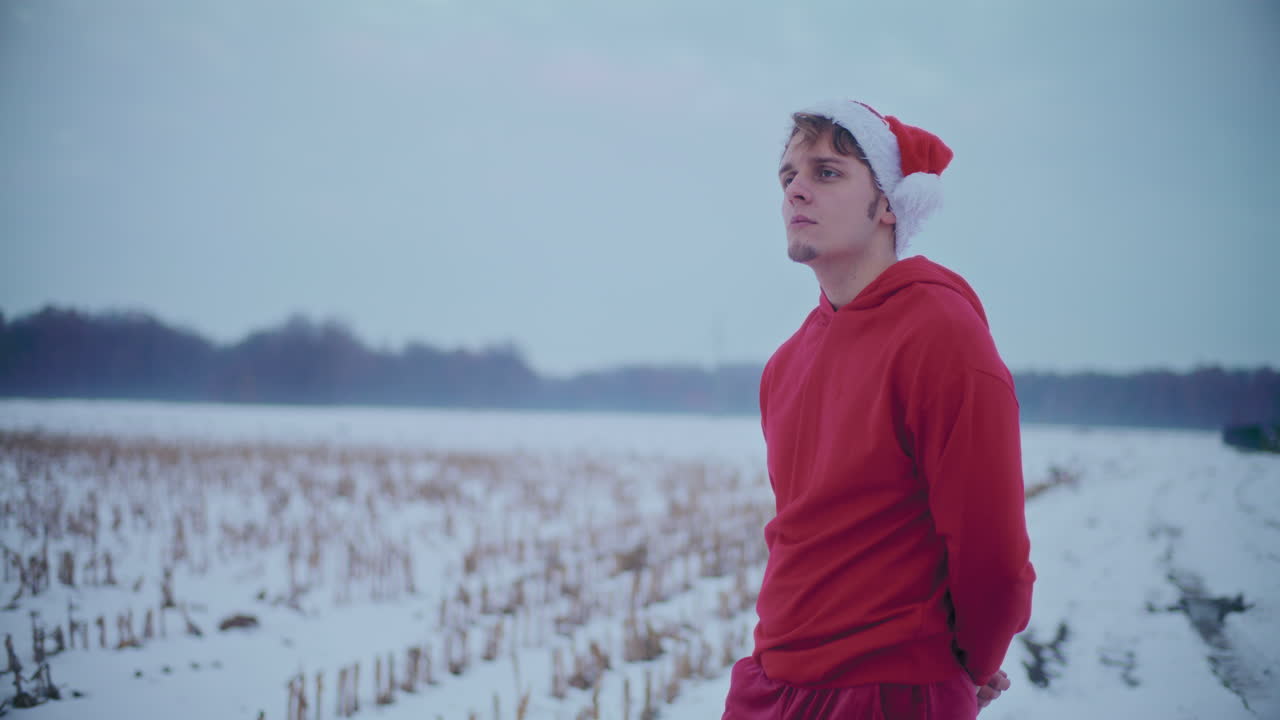 hombre pensativo con chaqueta roja con capucha y sombrero de santa de pie en el paisaje cubierto de nieve