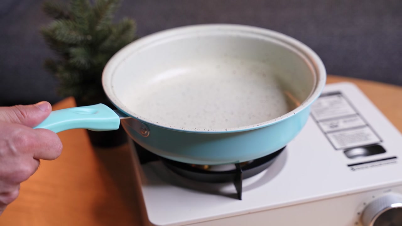 Primo piano di una mano che tiene una padella azzurra sopra un fornello a gas portatile acceso. La padella vuota si scalda su un tavolo di legno in un ambiente domestico moderno e interno con una piccola pianta.