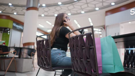 young student seated in a mall, engaging in a phone call while relaxing hand on table; blurred urban backdrop with shop inscriptions