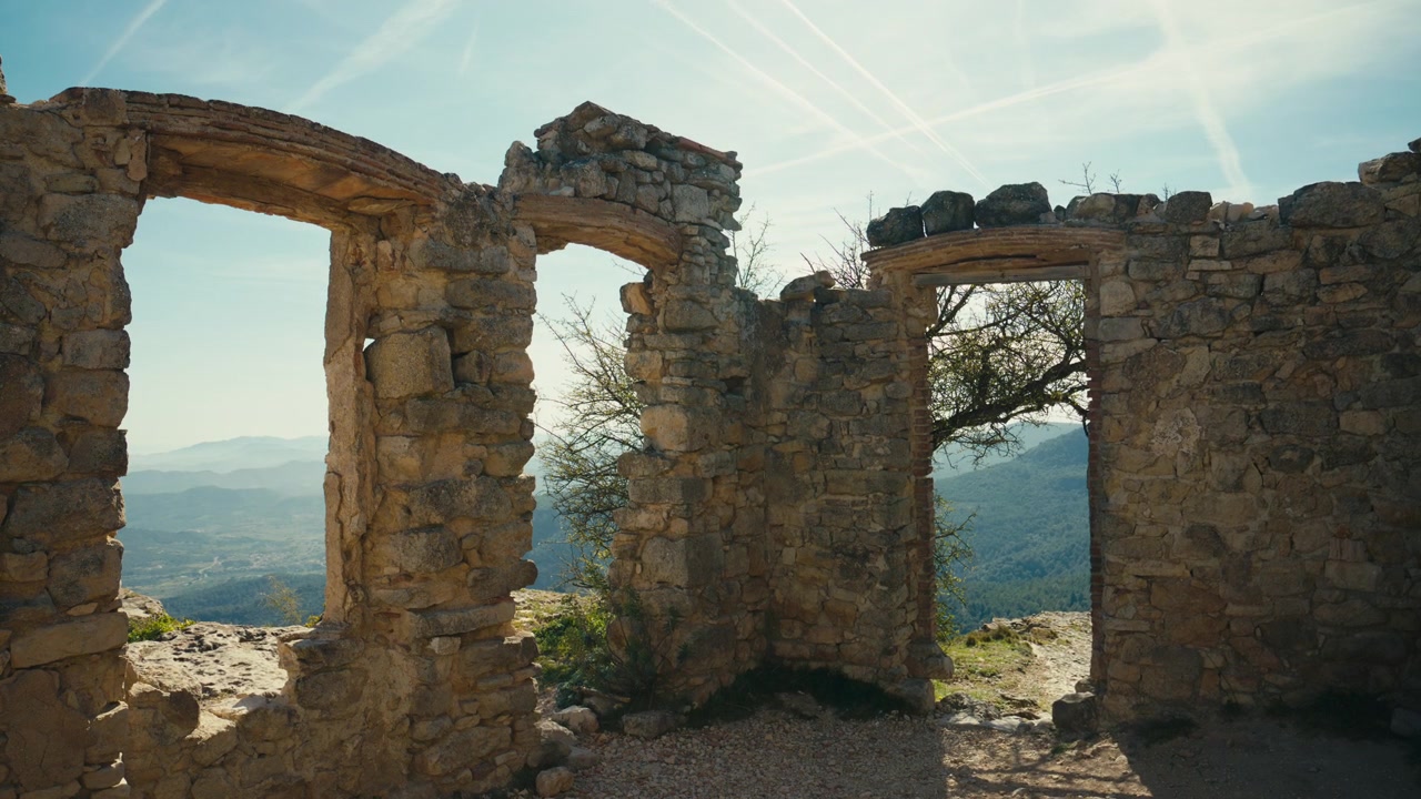 Aperture in pietra consumata in una struttura in rovina si affacciano sulle montagne della Catalogna, Spagna, dove il cielo luminoso, la muratura grezza e le creste distanti creano una drammatica scena storica.
