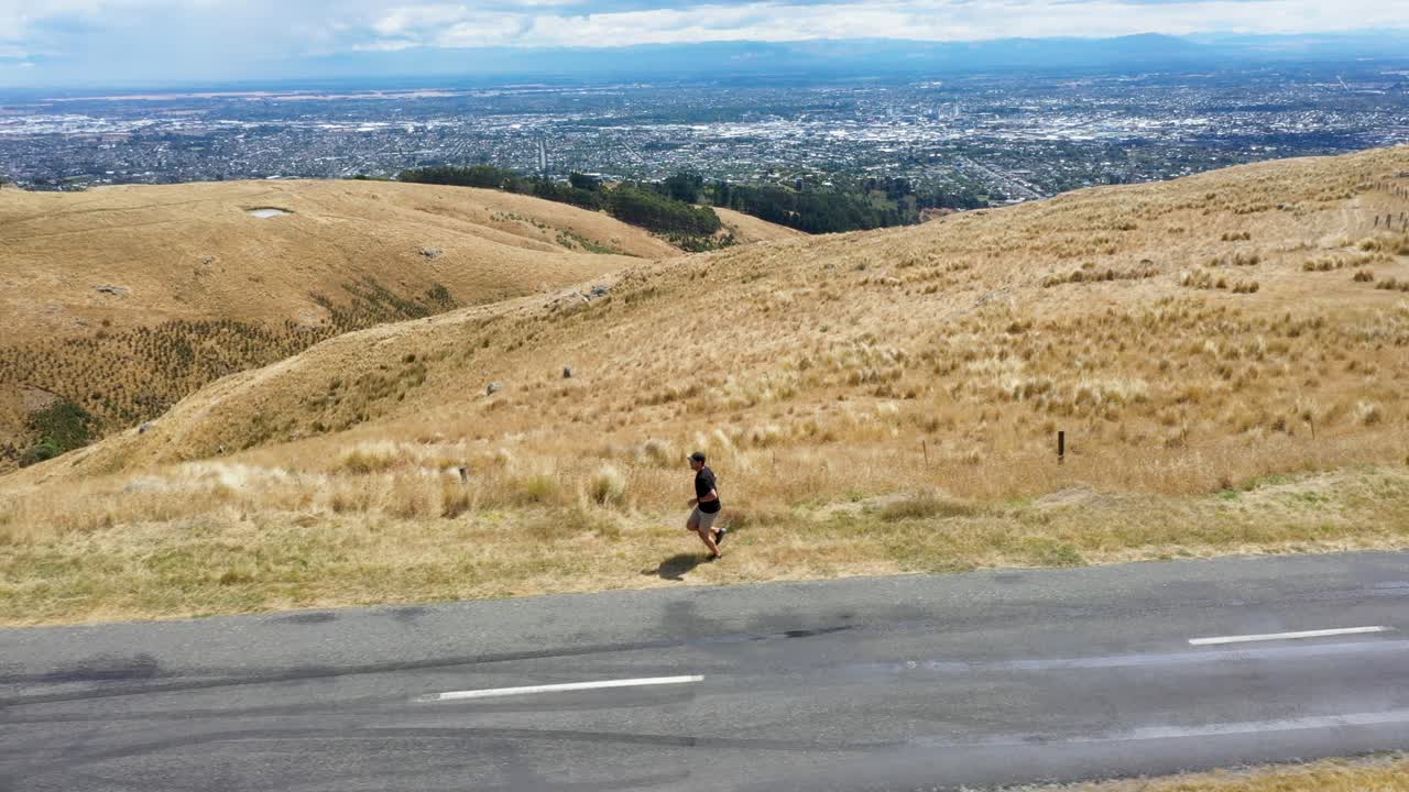Profile tracking shot of man running on grass by road on hill.