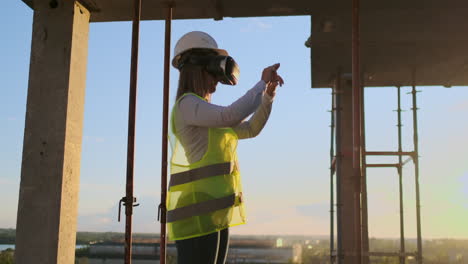 woman inspector in vr glasses and helmet checks the progress of the construction of a skyscraper moving his hands at sunset visualizing the plan of the building