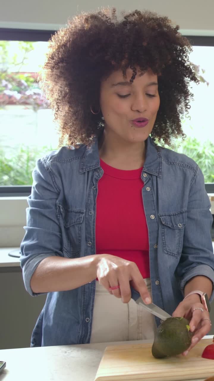 Video verticale: Lei, vestita in denim, posiziona un avocado su un bancone vicino a una finestra, lo taglia con un coltello e rimuove il nocciolo per mangiarlo.