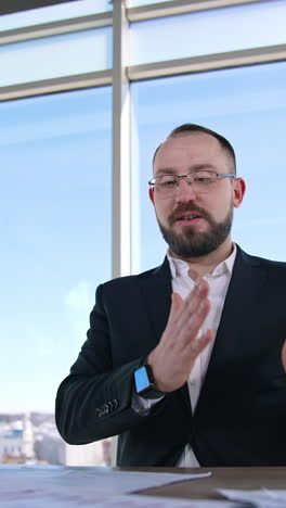 Successful businessman leading a conference. Male entrepreneur in glasses and costume sitting at the table and talking to someone in light office. Vertical video