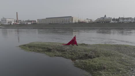 Toma-Aérea-De-Un-Dron-De-Una-Mujer-Con-Un-Vestido-Rojo-Brillante-En-Una-Pequeña-Isla-Cerca-De-La-Ciudad