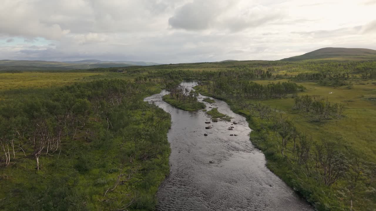 Um rio tranquilo serpenteia por uma tundra baixa e arbustos esparsos sob um céu aberto, ao longo da rodovia E69 em Finnmark, Noruega – Drone voando para frente