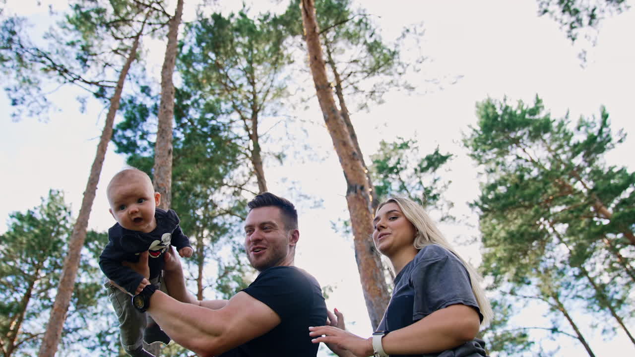 Smiling bearded man raises newborn son up. Blonde woman standing behind her husband. Low angle view. Nature backdrop.