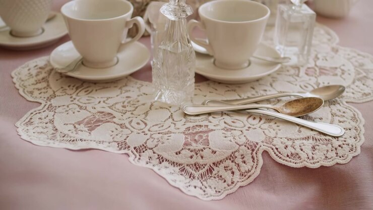 Close detail of stacked metal teaspoons resting diagonally on ornate round lace doily over pink textured tablecloth while faceted clear glass shaker stands between white teacups, soft warm light