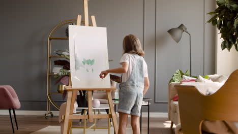 rear view of a blonde girl painting a plant with a brush on a lectern in the living room at home 2