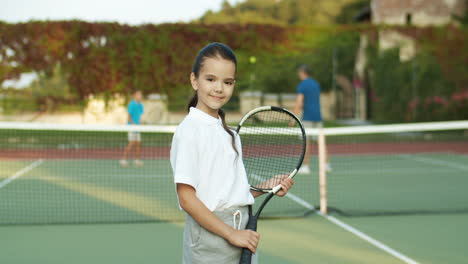 Portrait-Of-Cute-Little-Girl-Holding-Racket-And-Smiling-Cheerfully-At-The-Camera-While-Standing-On-An-Outdoor-Tennis-Court