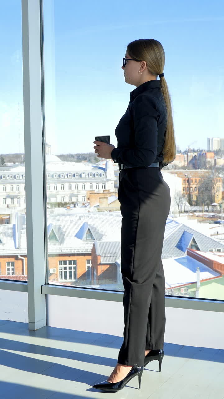 Beautiful business woman standing with coffee. Full-length portrait of an elegant woman near the office window. Panoramic window view with a city background. Vertical video