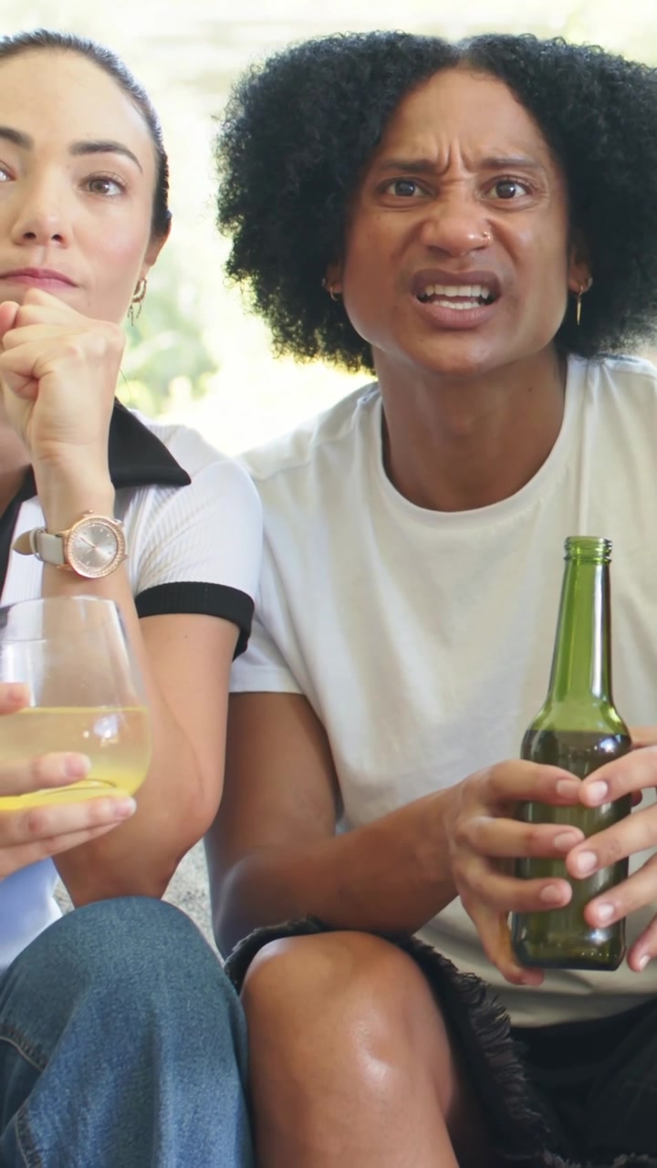 Vertikales Video: Reagierender Mann in weißem T-Shirt hält grünes Bier auf der Couch, schaut mit einem Glas zur Seite.