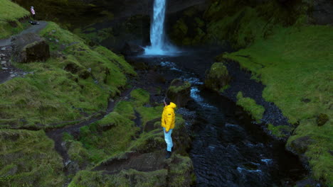 Man-In-Yellow-Jacket-Stands-On-Rock-In-Front-Of-Kvernufoss-Waterfall-In-Iceland