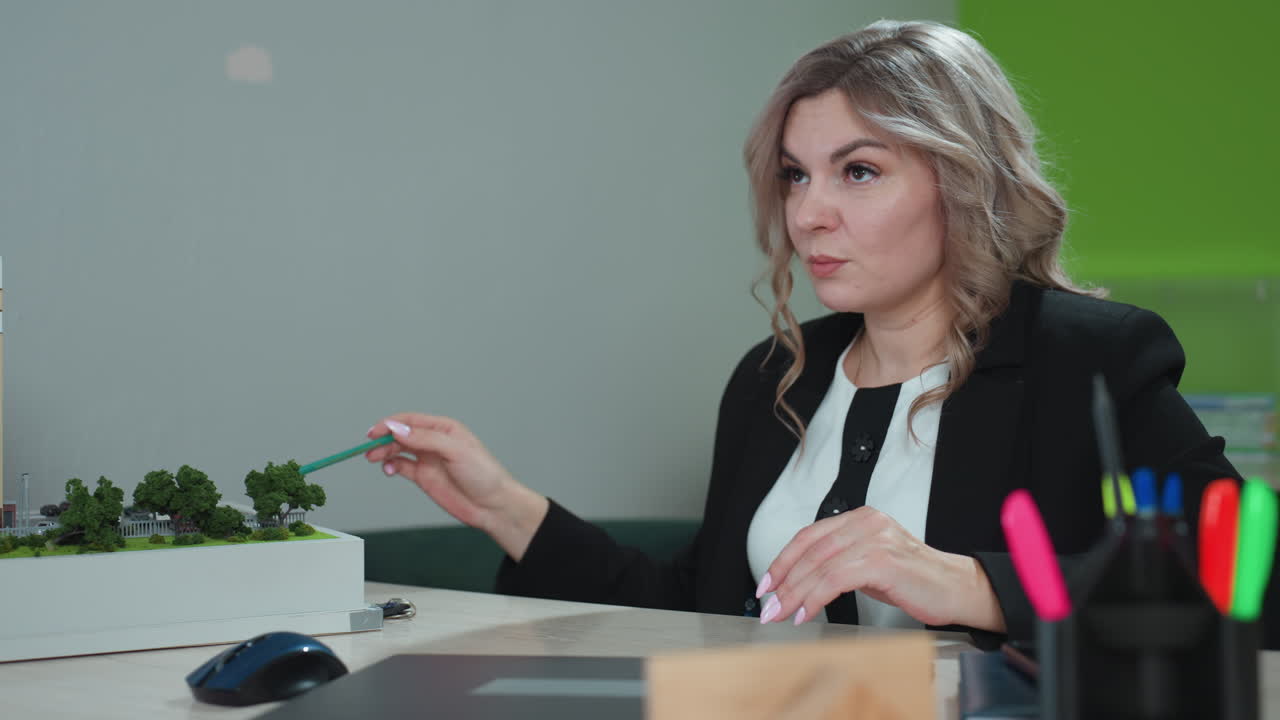 realtor explaining building miniature to client using pencil while seated at office desk with colorful pens in holder and architectural model in foreground
