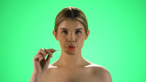 medio disparo estático de una joven mujer bonita sosteniendo una deliciosa piruleta en forma de corazón en la cámara antes de lamerla y disfrutar de la dulzura frente al fondo verde en cámara lenta
