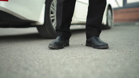 close-up leg view of individual standing near white car in urban environment, person's legs positioned beside vehicle, emphasizing modern car and casual stance in outdoor setting