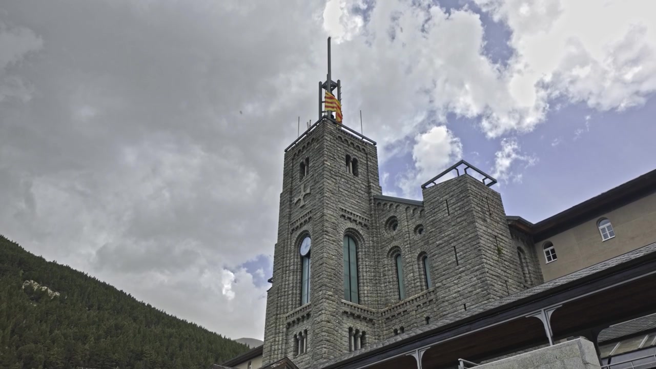 Maestoso campanile in pietra del Santuario di Nuria, situato nei Pirenei della Catalogna, con la bandiera catalana che sventola contro un cielo drammatico pieno di nuvole in rapido movimento.