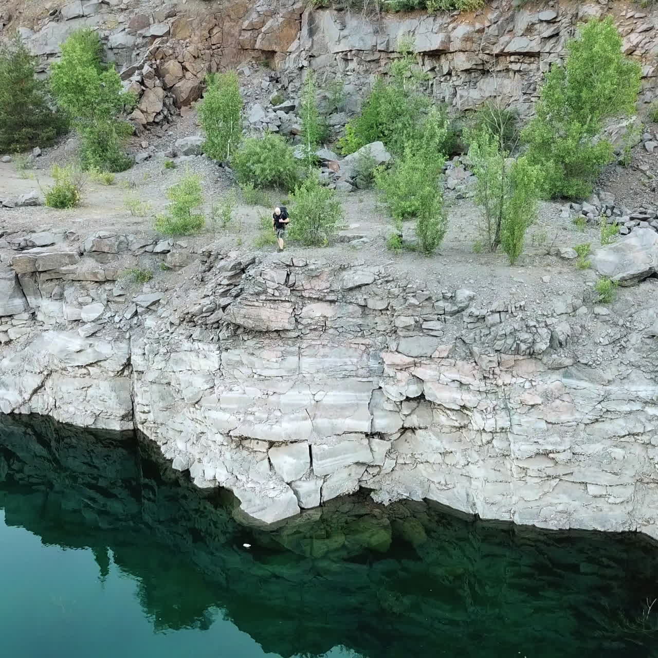 Rocky cliff reflects in green water and tourist in summer clothes looking into it. Natural landscape in the mountains outdoors. Camera moves bottom top.