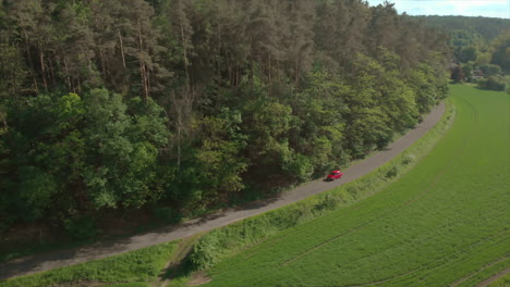 Coche-Rojo-Conduciendo-Por-Una-Carretera-Recta-A-Lo-Largo-Del-Bosque-Y-El-Campo-Verde,-Tiro-De-Drones